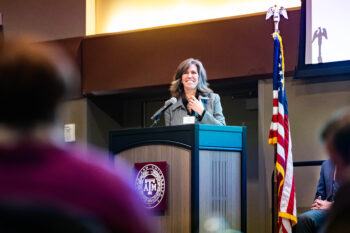 A woman addressing an audience from a lectern adorned with the Texas A&M University seal.