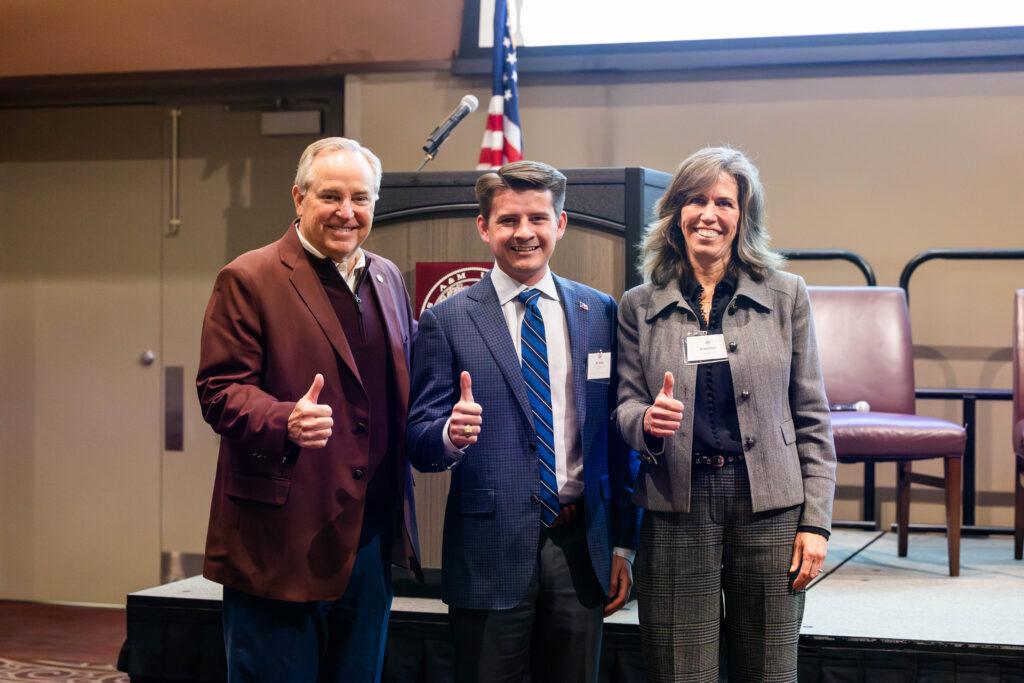 A photo featuring three individuals dressed in business attire giving the Texas A&M 'Gig'em' thumbs-up.