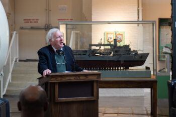 An image of a man addressing an audience from a lectern with a ship model positioned behind him.