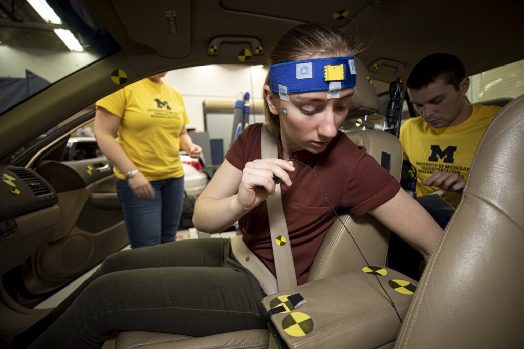 A woman, with a sensor strip on her forehead, secures herself in the passenger seat of a test vehicle while a researcher in the back seat operates a laptop.