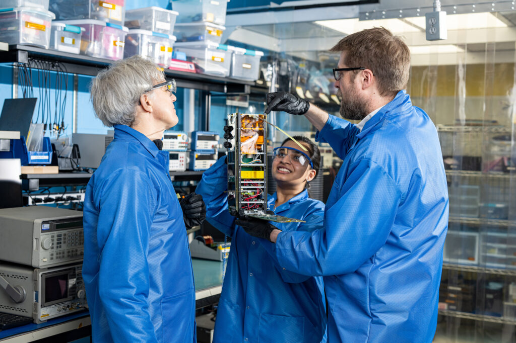 Three researchers clad in blue lab coats and black gloves are conversing in a group while inspecting a compact, boxy satellite held by the researcher in the center. The satellite measures about a foot in height and four inches in width and depth.