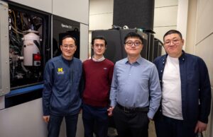 Four men stand by a window displaying a cylindrical structure along with various wires and tubes.