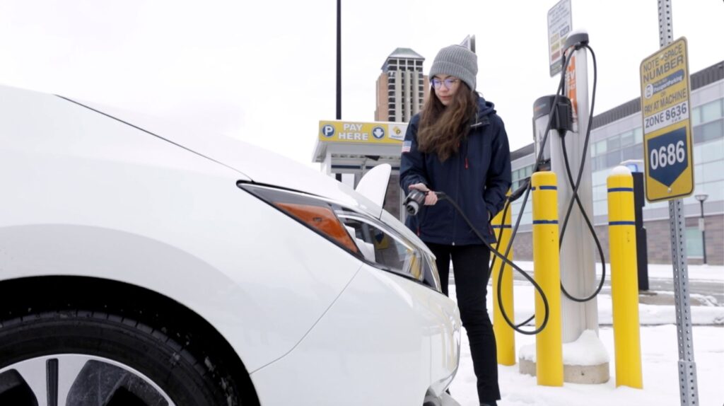 A woman, donned in a jacket and winter hat, gets ready to connect an electric vehicle to a charger.