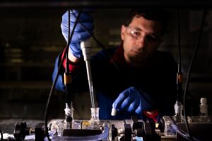 A scientist wearing a blue lab coat and nitrile gloves places a pipette into a cylindrical, glass chamber via a narrow spigot on its top.