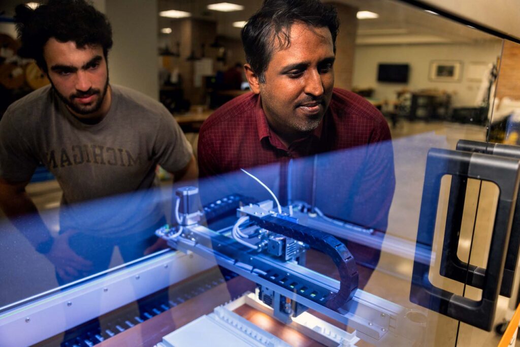 Two individuals are reflected in the glass of the 3D printer, which is fabricating a plastic structure.
