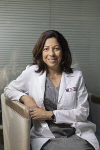 A woman in a white medical coat with a UGA logo smiles while seated and facing the camera.
