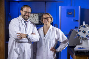 Two medical researchers in white coats pose in a lab illuminated with blue light.