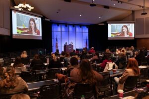 Audience members in an auditorium look up at two display screens during a children's literature conference. There is a stage between the screens.