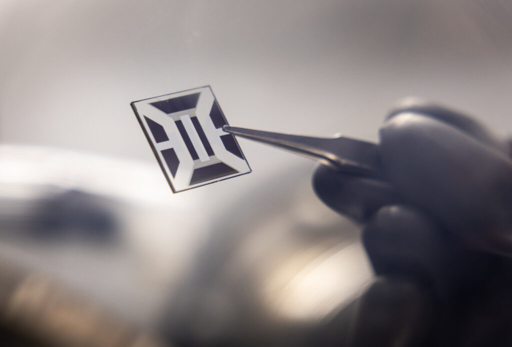 A transparent square, measuring a couple of centimeters per side, adorned with black markings, is grasped by tweezers.