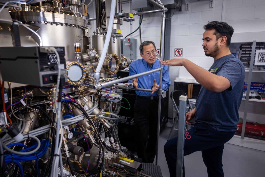 Two individuals are engaged in conversation, standing next to a large machine made of silver and gold with numerous tubes and valves connected.