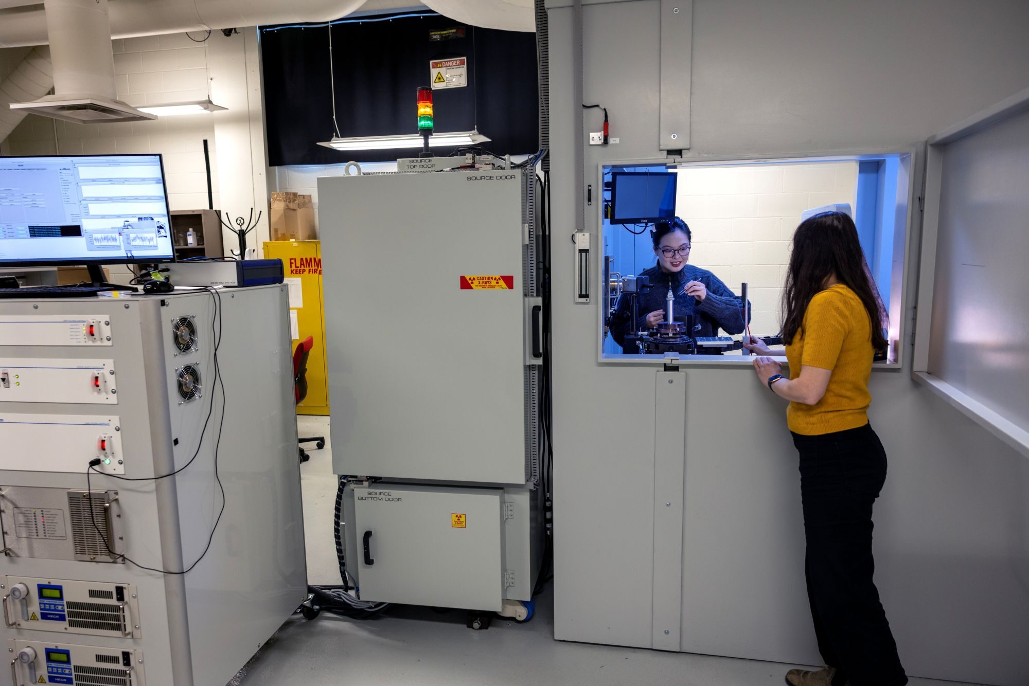 Two individuals operate in a lab: In the background, an open window reveals a small room where one woman adjusts a sample on a microscope holder. In the foreground, a second woman stands in the larger room, observing through the window. Two refrigerator-sized devices are situated to the left of the room with the microscope stand.