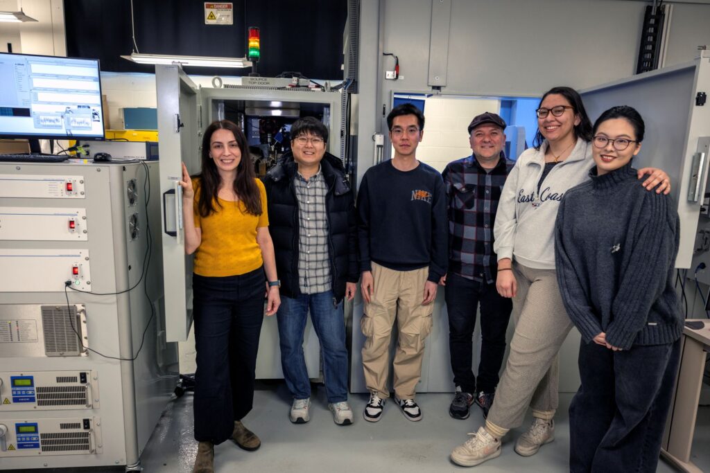 Six individuals stand next to each other in a lab in front of a large apparatus resembling a 5-foot-tall refrigerator with microscope apparatus inside the top drawer.