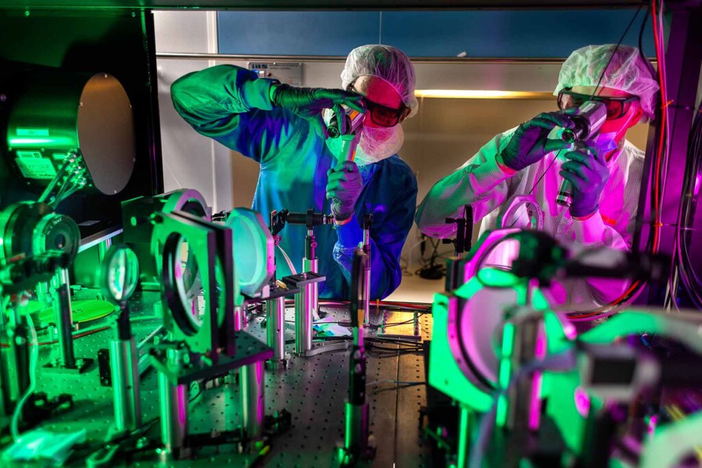 Two men in hairnets, beard nets, lab coats, gloves and protective eyeglasses peer through spyglass-like devices at the optics inside the laser cabinet, mostly circular and transparent devices mounted in metal stands.