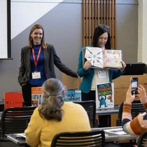 Two women at a literature conference hold up a picture book and talk to people during a panel.