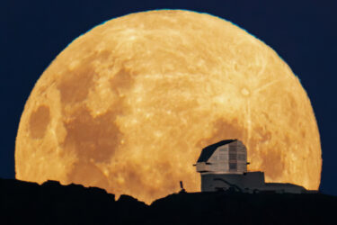 A large observatory positioned on a ridge with the full moon rising in the background
