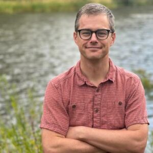 A man stands by a body of water with folded arms.