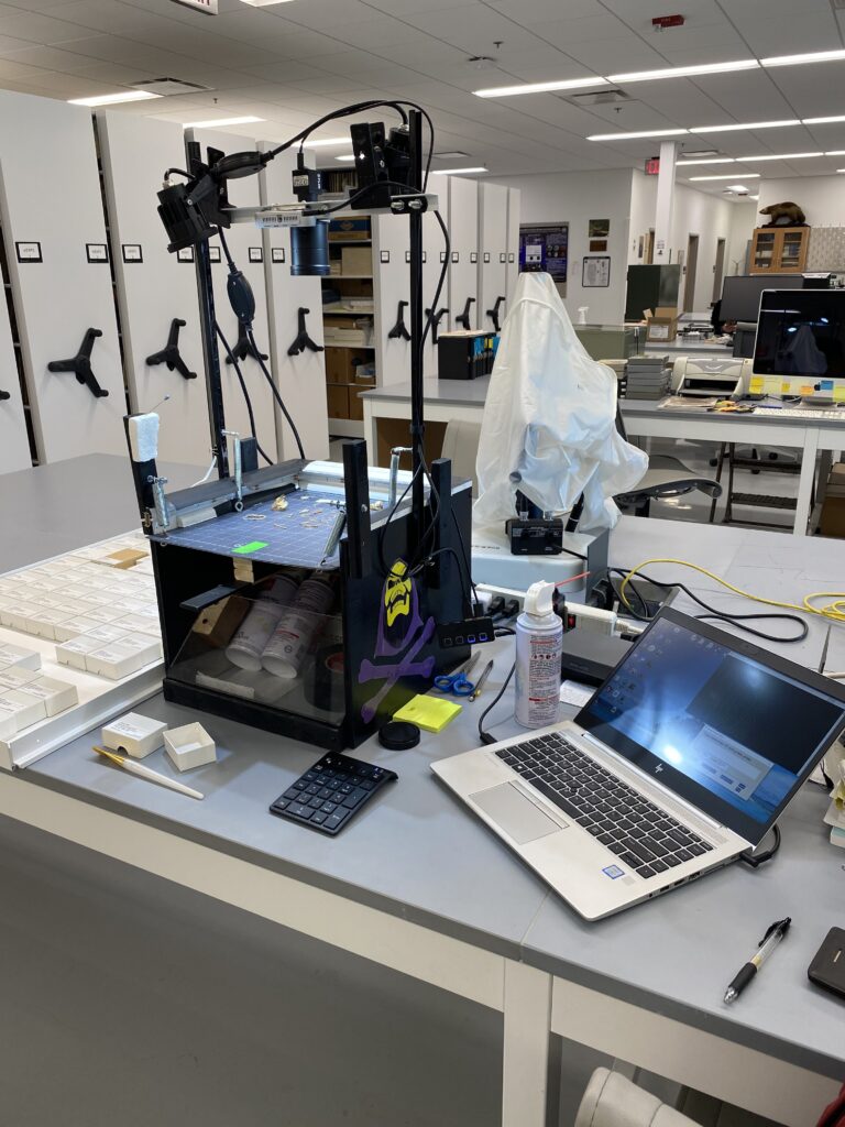 The Skelevision system is arranged on a table, before rows of specimen cabinets, consisting of a box-like platform for observing samples. A camera is positioned above the platform, directed at the specimens, and the system connects to a laptop.