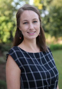 A brunette woman in a checked dress poses outdoors for an environmental portrait.