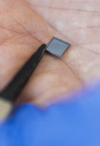 A close up of a hand holding a square silicone patch, just a few millimeters wide, embedded with tiny microneedles on one side. Tweezers maintain the patch in place.
