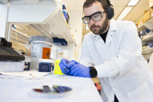 A man in a lab coat, goggles, and gloves uses tweezers to place a small silicone patch featuring microneedles into a petri dish in a laboratory environment.
