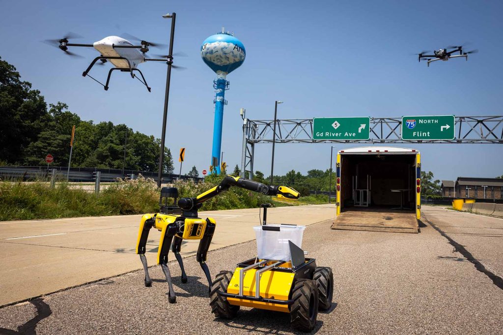 On Mcity's mock freeway, a four-legged, long-necked robot, about the height of an average dog at the shoulder, is next to a shorter rugged wheeled robot carrying a plastic bin. Above, a white cargo drone with a dog-sized body and smaller quadcopter fly.