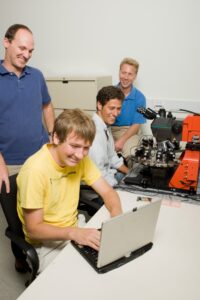A man in a yellow shirt operates a computer while another is at a device resembling a complex microscope. Two additional men in blue shirts observe behind them.