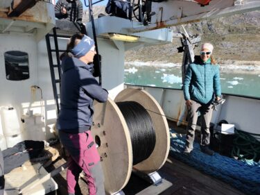 A large cable drum is being unwound by one individual. Another person stands in the background.