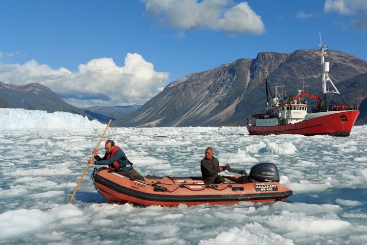 A compact inflatable and motorized boat navigates through ice-laden waters. A larger vessel is anchored in front of a glacier behind.