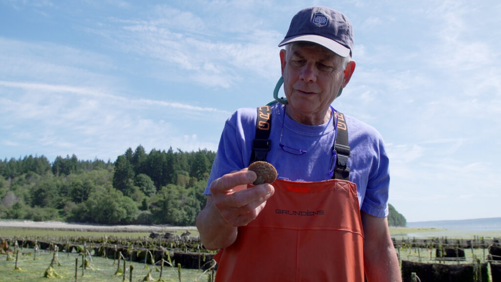 A man in vivid orange waders holds a small clam.
