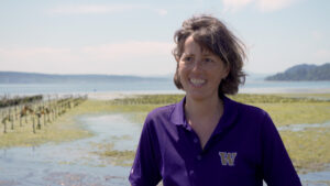 A woman stands in tidelands covered in seaweed. She is dressed in a purple polo shirt featuring the UW logo.