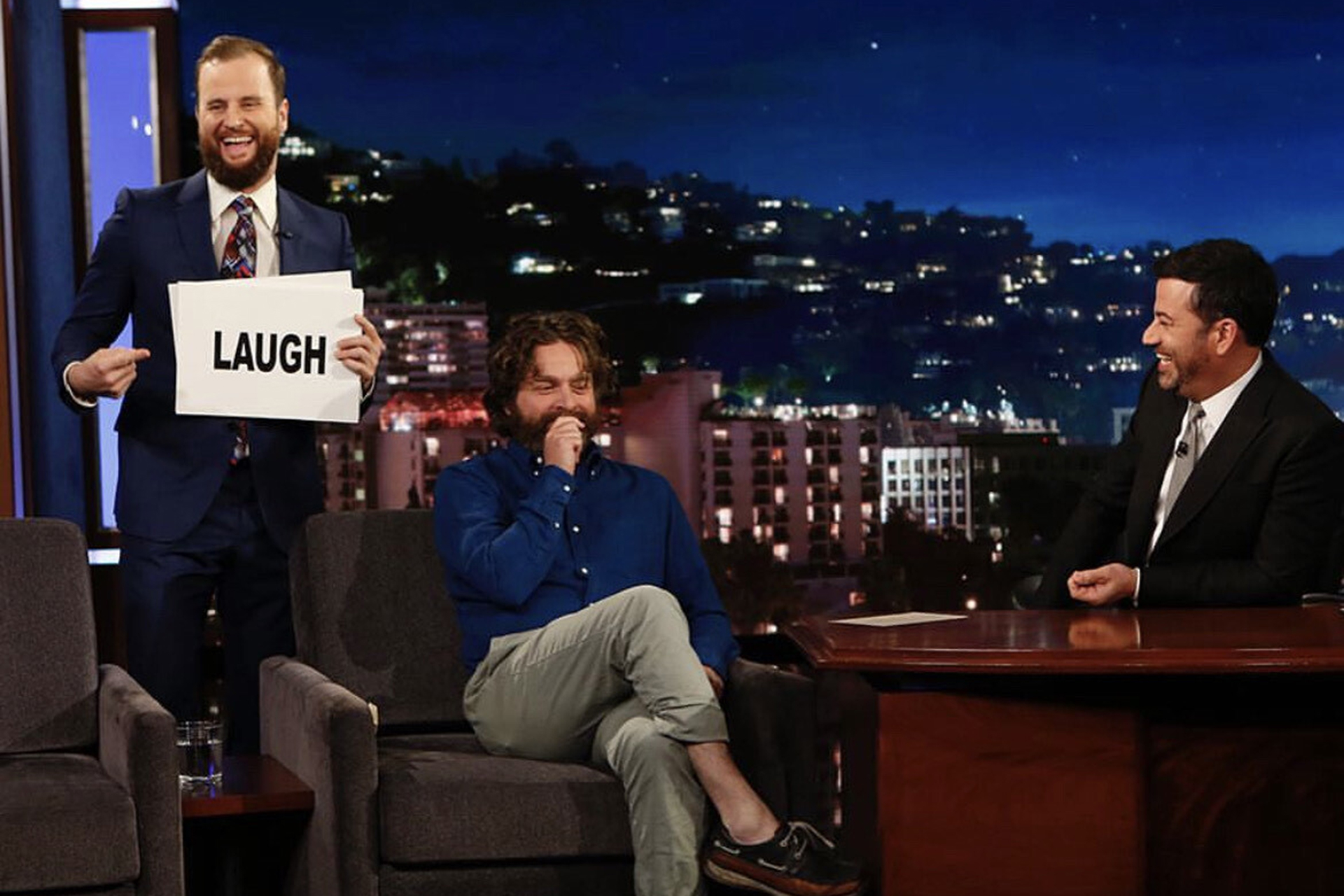 Will Burke displays a "laugh" sign as Zach Galifianakis converses with Jimmy Kimmel.