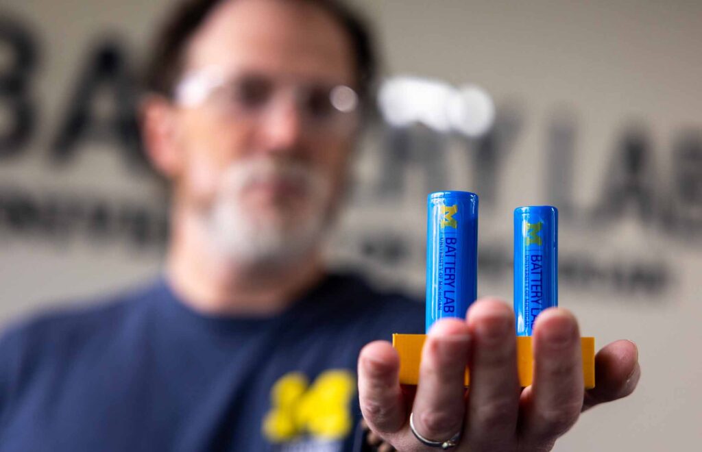 A man holds two blue cylindrical batteries on an orange platform.