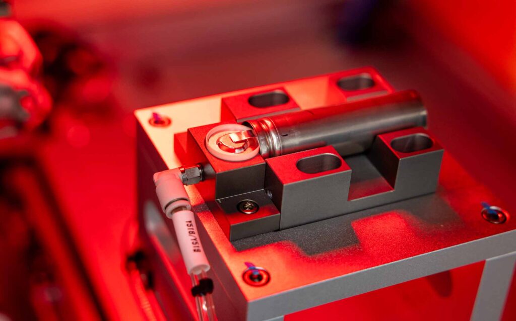 Cylindrical battery in a holder on a laser welding machine with red lighting.