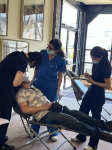 Three dental practitioners in scrubs examine a patient, who reclines on a chair.