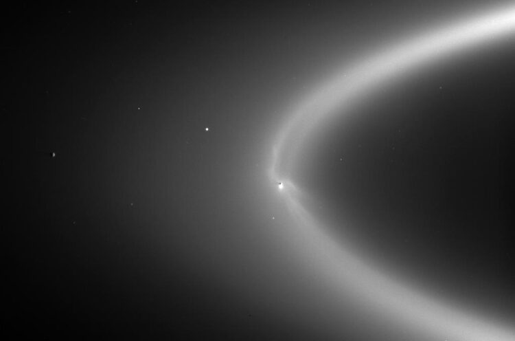 A monochromatic image demonstrating a bright white ring of ice in the cosmos. Saturn’s moon Enceladus is represented as a tiny black dot within the ring.