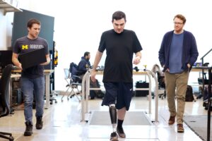 Three men walk through a lab with a treadmill set into the floor behind them. In the center, Seelhoff wears a prosthetic leg that integrates its electronics within a slim calf-shaped casing. On the left, Kevin carries an open laptop computer.