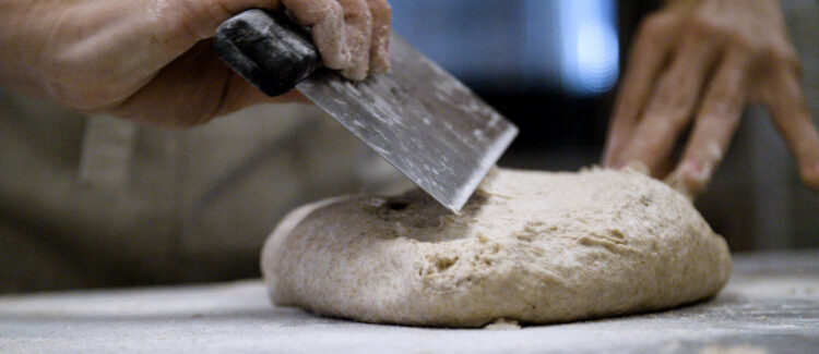 An individual utilizes a dough scraper to manipulate a mass of bread dough.